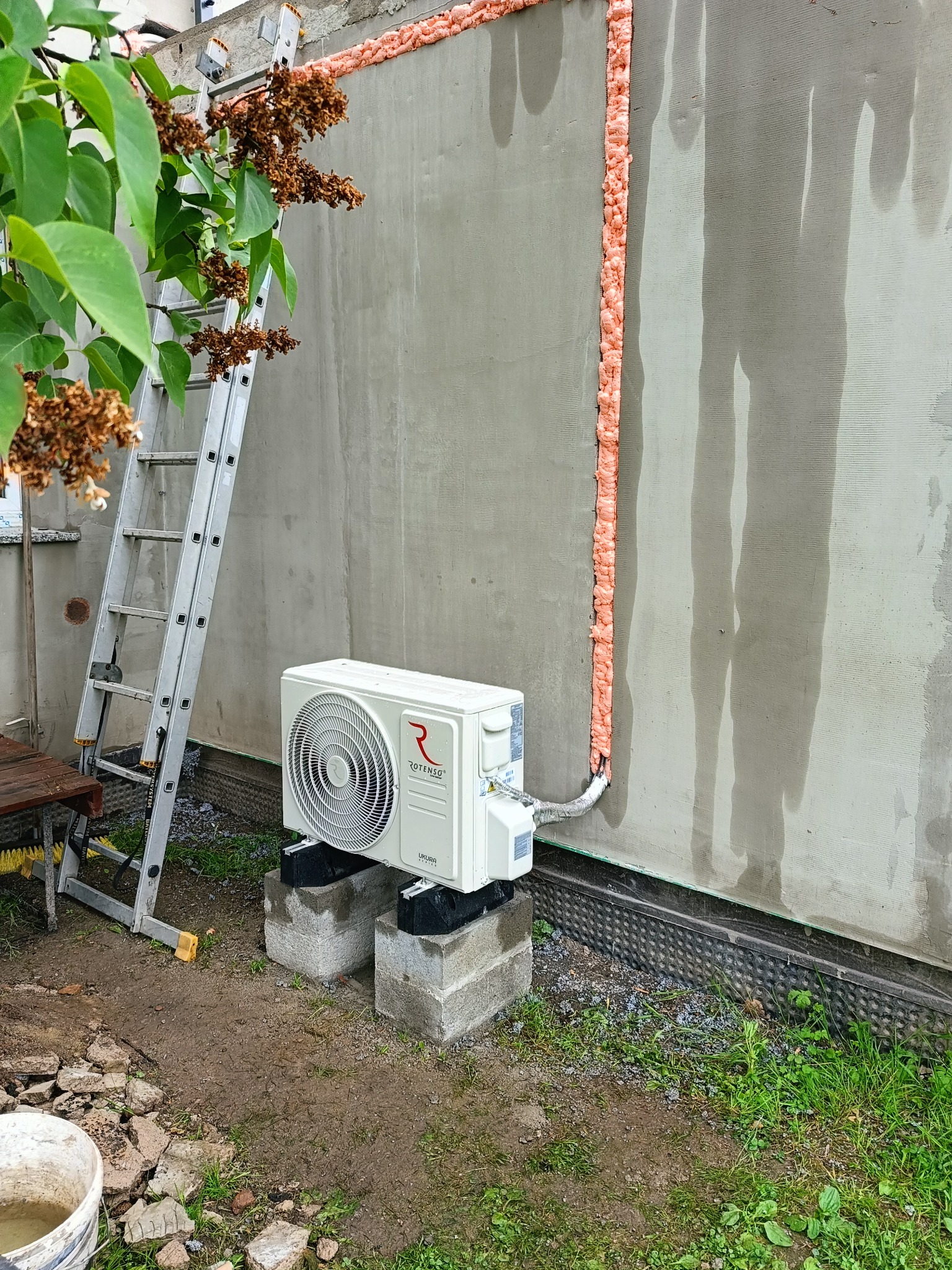 Biały klimatyzator Rotenso na betonowych blokach przy ścianie budynku z pionową linią pianki montażowej, drabina oparta o ścianę, widok z zewnątrz.