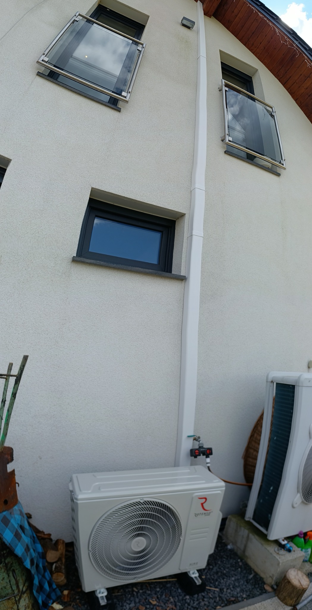 Biała jednostka zewnętrzna pompy ciepła Rotenso, zamontowana przy ścianie budynku z oknami balkonowymi. Widoczne elementy instalacji i rura odprowadzająca.
