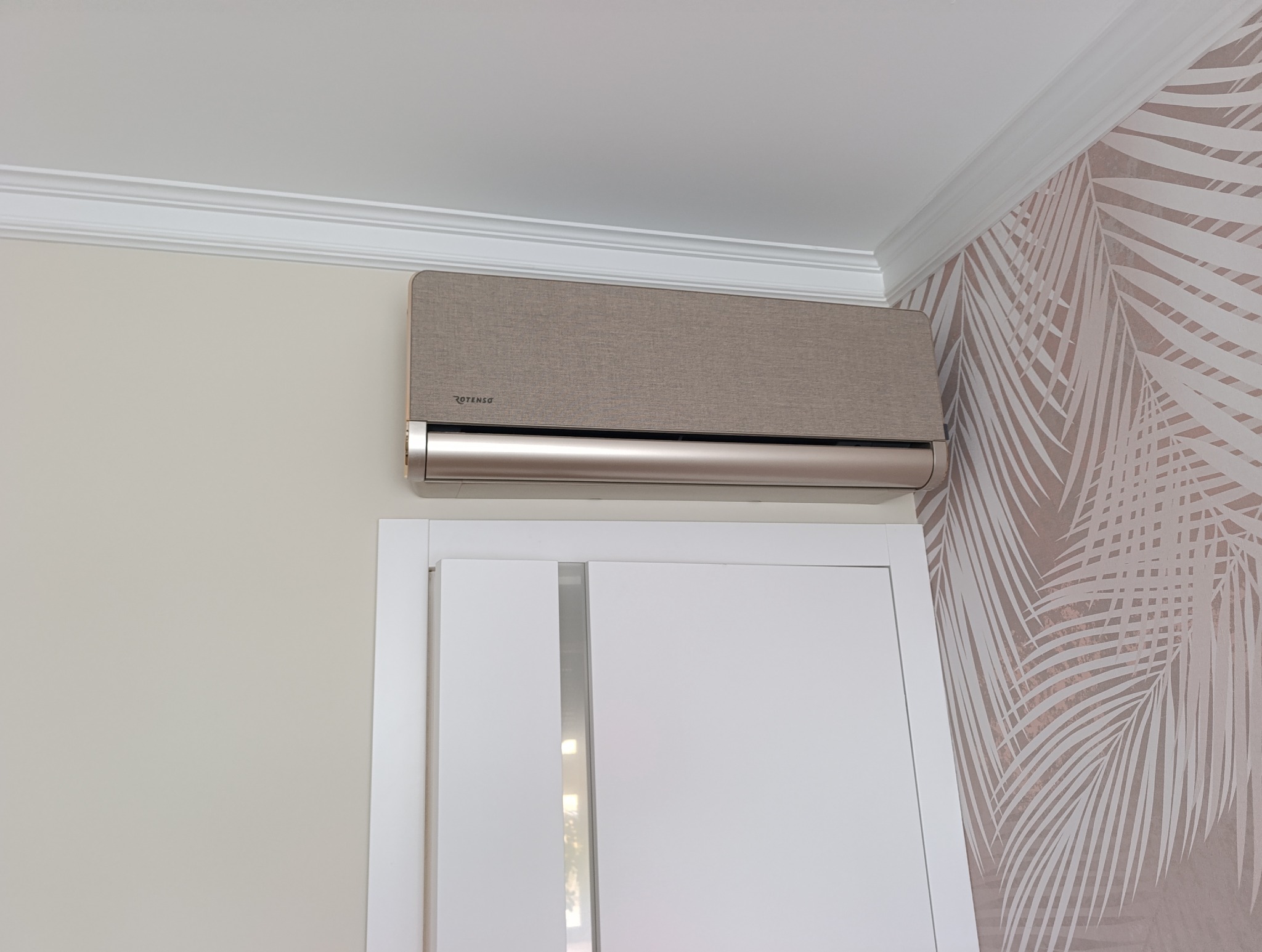 Beżowy klimatyzator Rotenso, zamontowany nad drzwiami w pokoju z tapetą w palmy, eleganckie wykończenie sufitu.