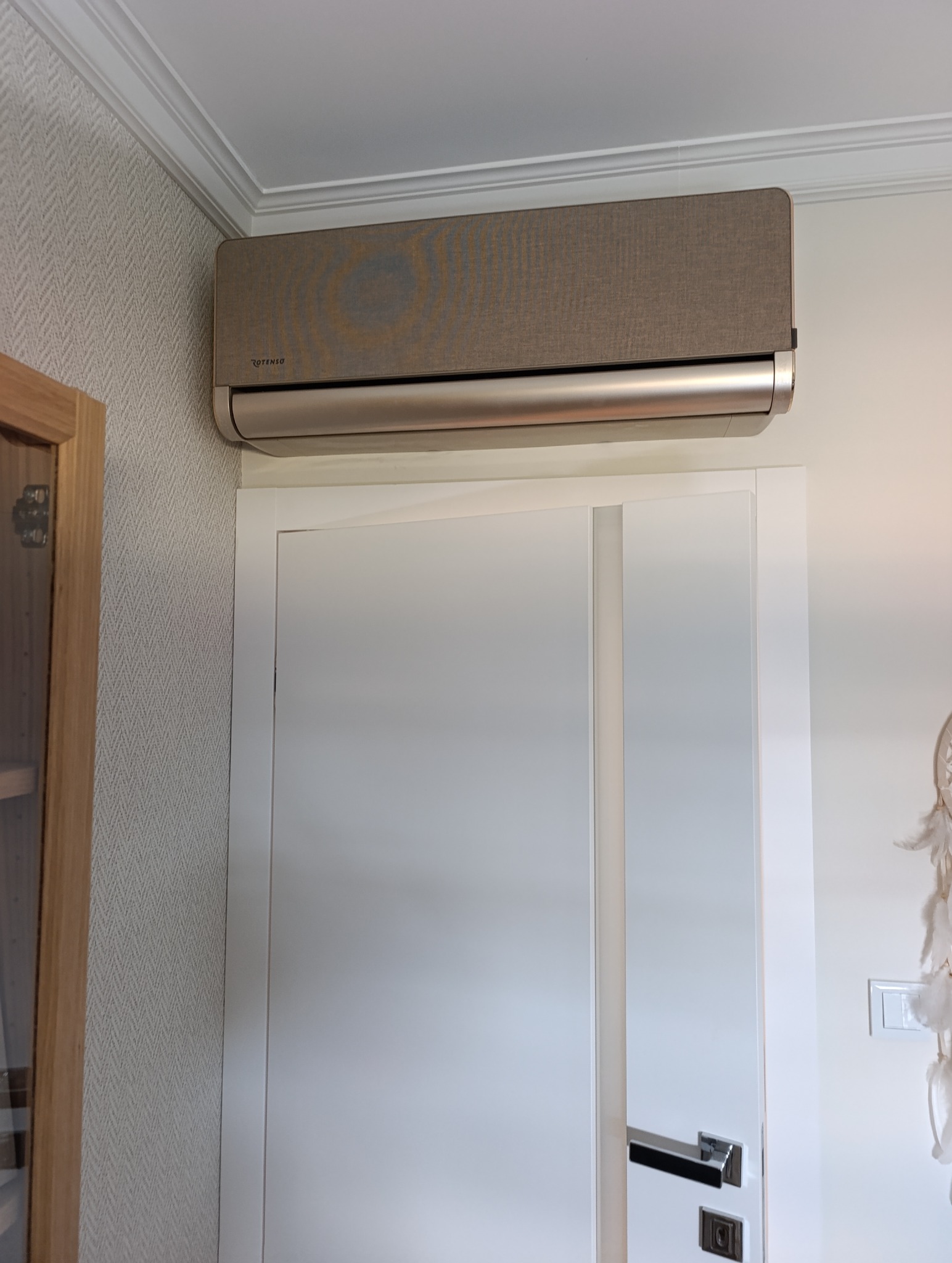 Beżowy klimatyzator Rotenso zamontowany nad białymi drzwiami w pomieszczeniu o jasnej ścianie z wzorem. Widoczny fragment drzwi i dekoracja na ścianie.