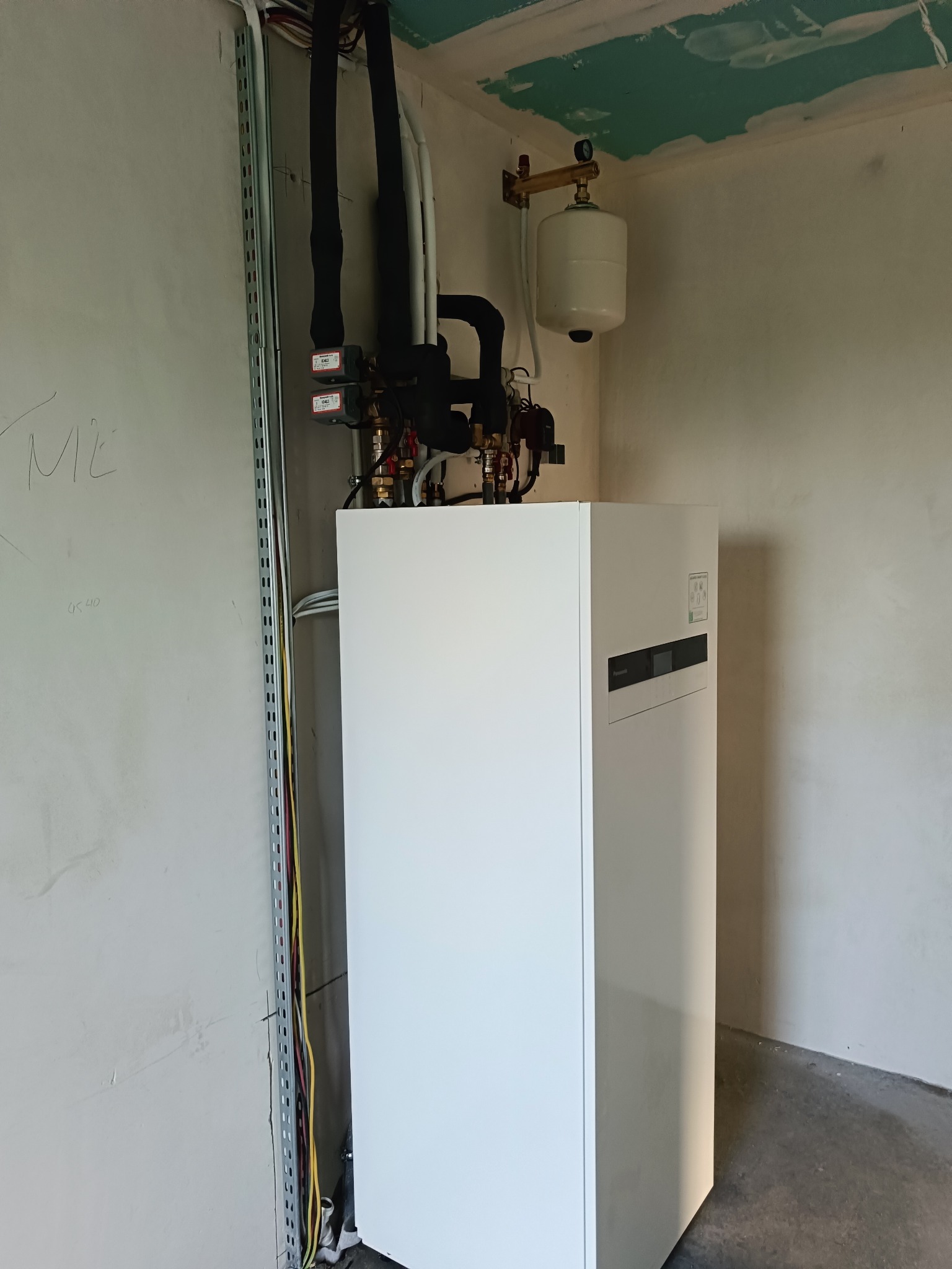 Biała pompa ciepła z czarnymi rurami i zbiornikiem wyrównawczym na ścianie w surowym wnętrzu, widoczne przewody elektryczne w korytku.