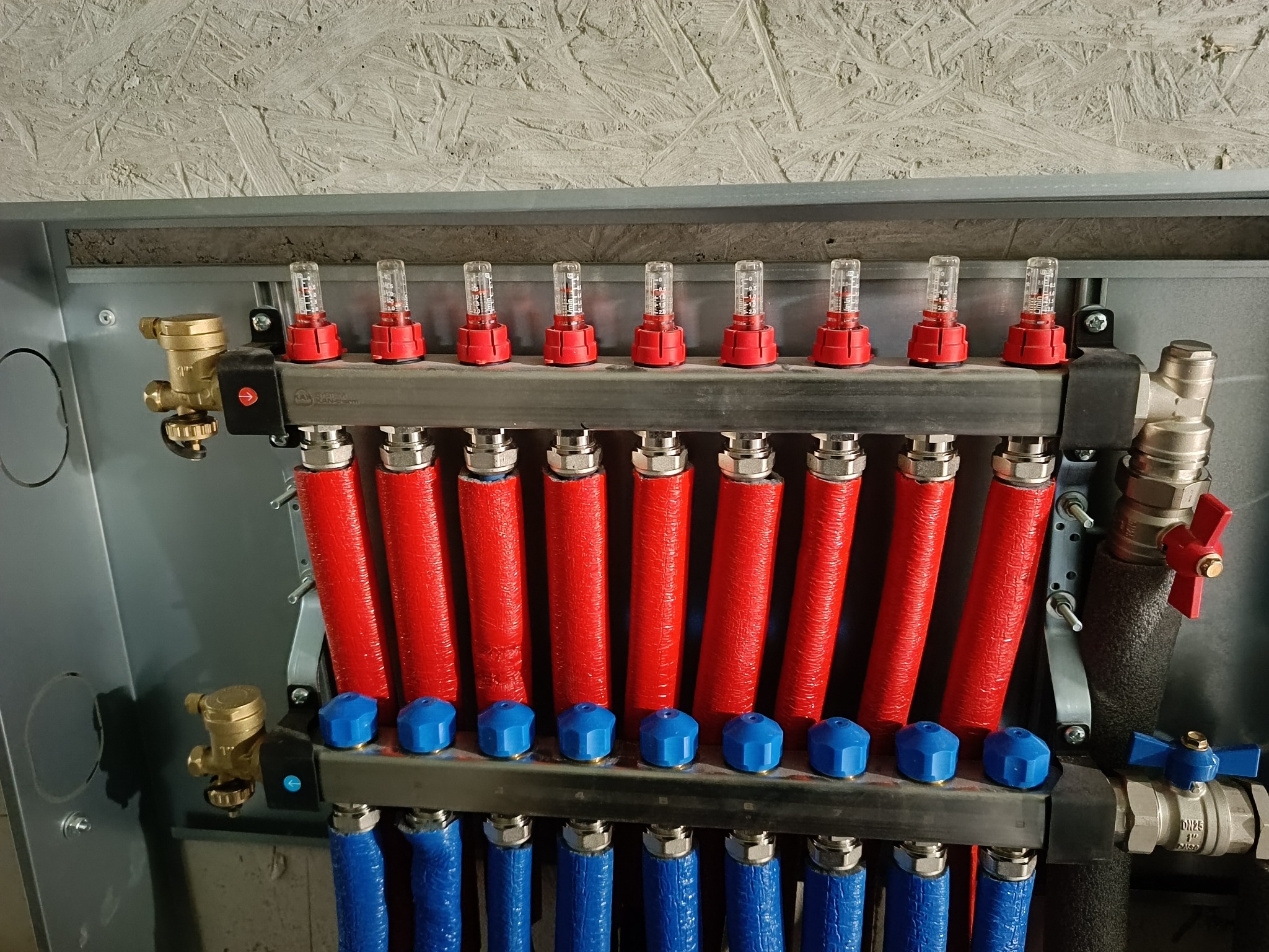 Rozdzielacz ogrzewania podłogowego z czerwonymi i niebieskimi rurami w metalowej obudowie, widoczne zawory i liczniki przepływu.