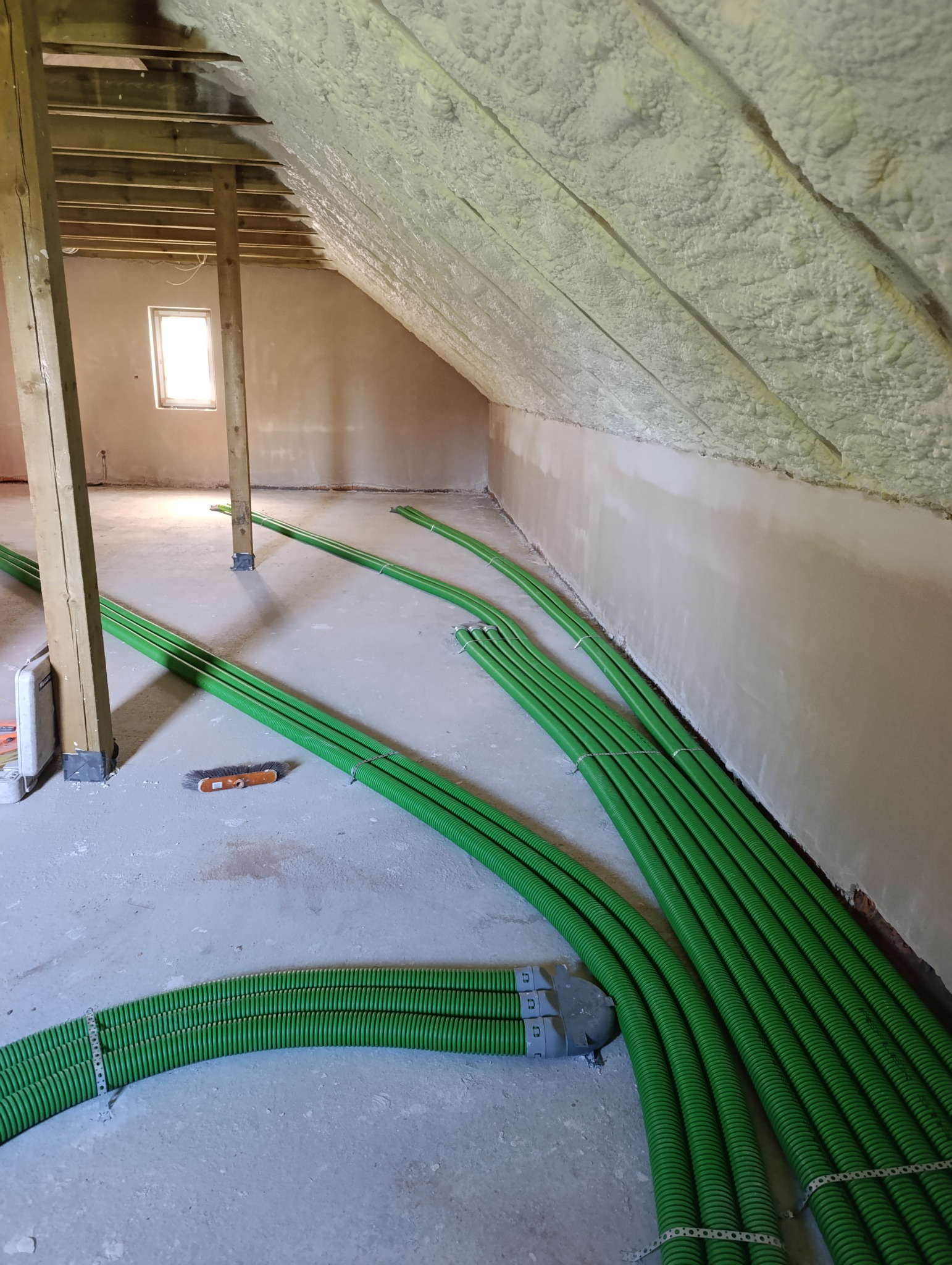 Zielone rury wentylacyjne ułożone na poddaszu z ocieplonym stropem, widoczne elementy mocujące. W tle okno i drewniane słupy konstrukcyjne.