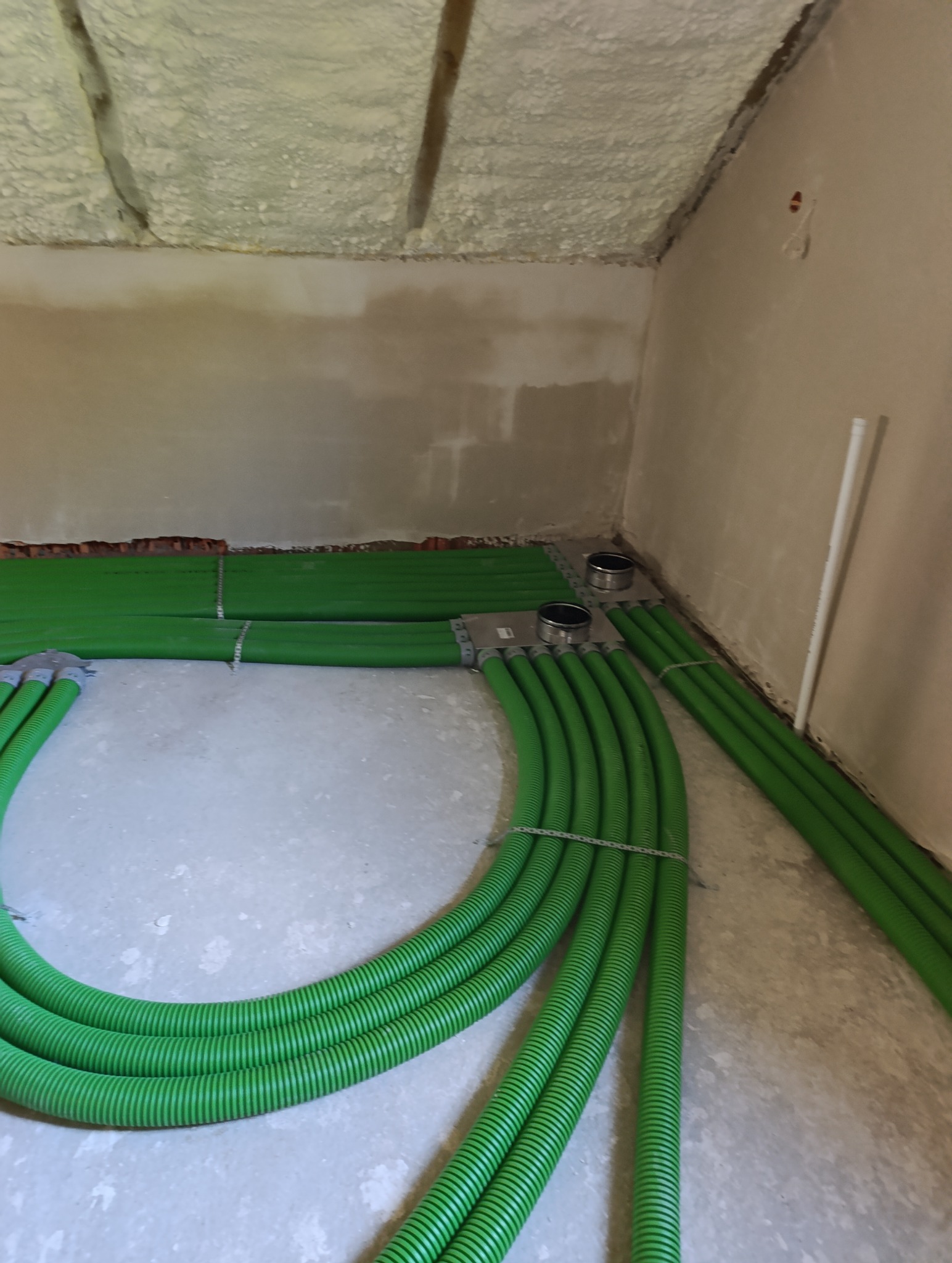 Zielone rury wentylacyjne ułożone na podłodze w surowym wnętrzu z pianką na suficie. Instalacja w trakcie realizacji, widoczne elementy montażowe.