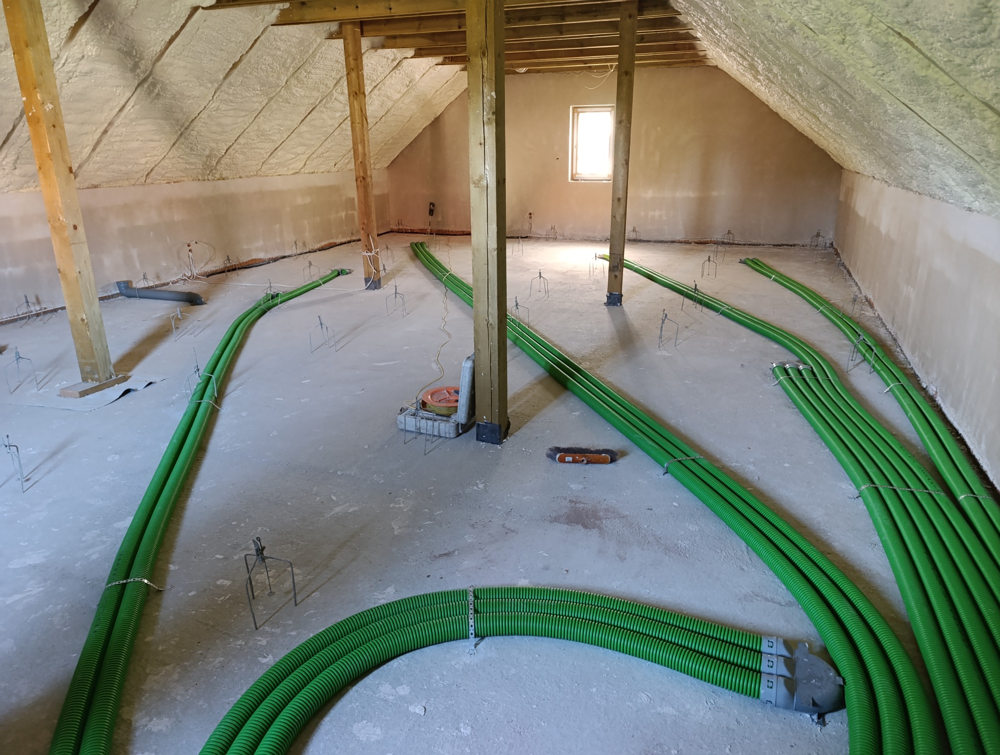 Montaż zielonych rur w podłodze na poddaszu, przygotowanie do wylania posadzki. Widoczne narzędzia i elementy mocujące.