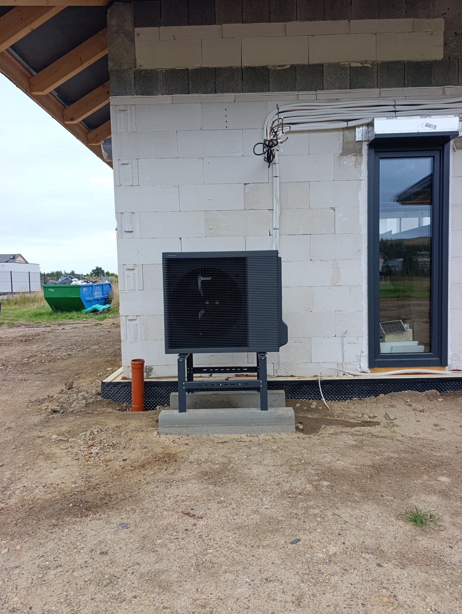 Czarna pompa ciepła Panasonic zamontowana na zewnątrz budynku z białego bloczka, widoczne rury instalacyjne i okno z ciemną ramą.