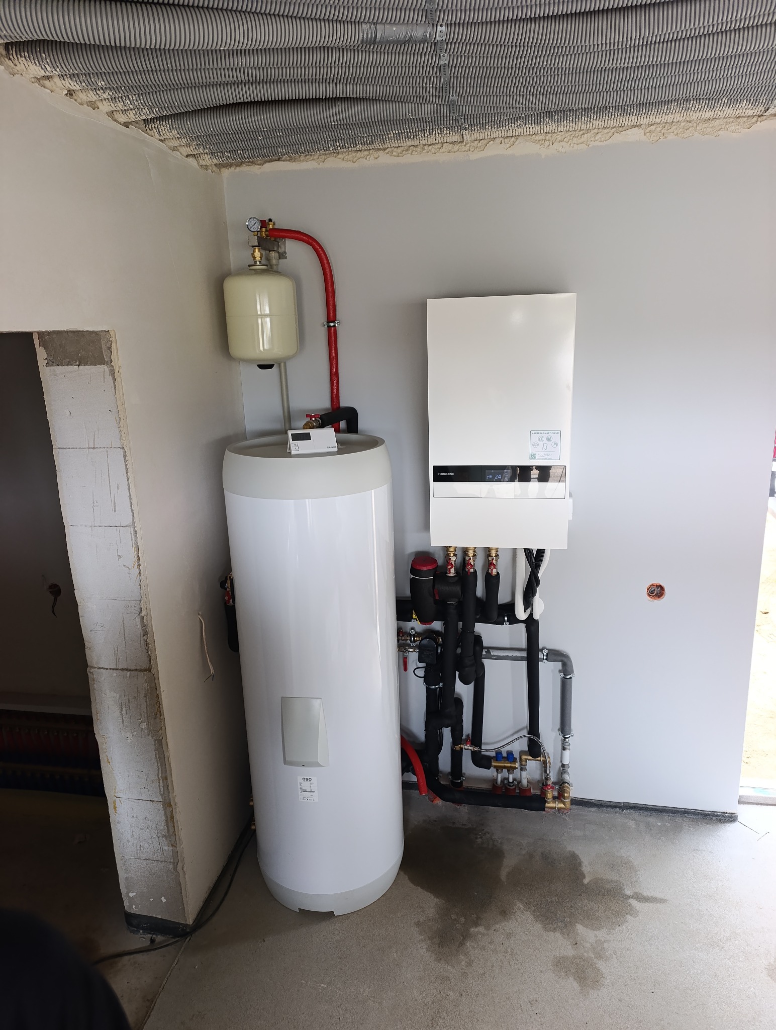 Biały zasobnik ciepłej wody użytkowej z podłączonymi rurami i panelem sterowania, zainstalowany w pomieszczeniu z widoczną instalacją wentylacyjną pod sufitem.