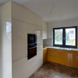 Jasna kuchnia z frontami w kolorze ecru i musztardowym, z zabudowaną piekarnikiem i widokiem na ogród przez okno, na brązowej podłodze.