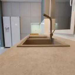 Beżowy, dwukomorowy zlewozmywak z baterią w nowoczesnej kuchni z szarymi szafkami i białym blatem, widok z perspektywy blatu.