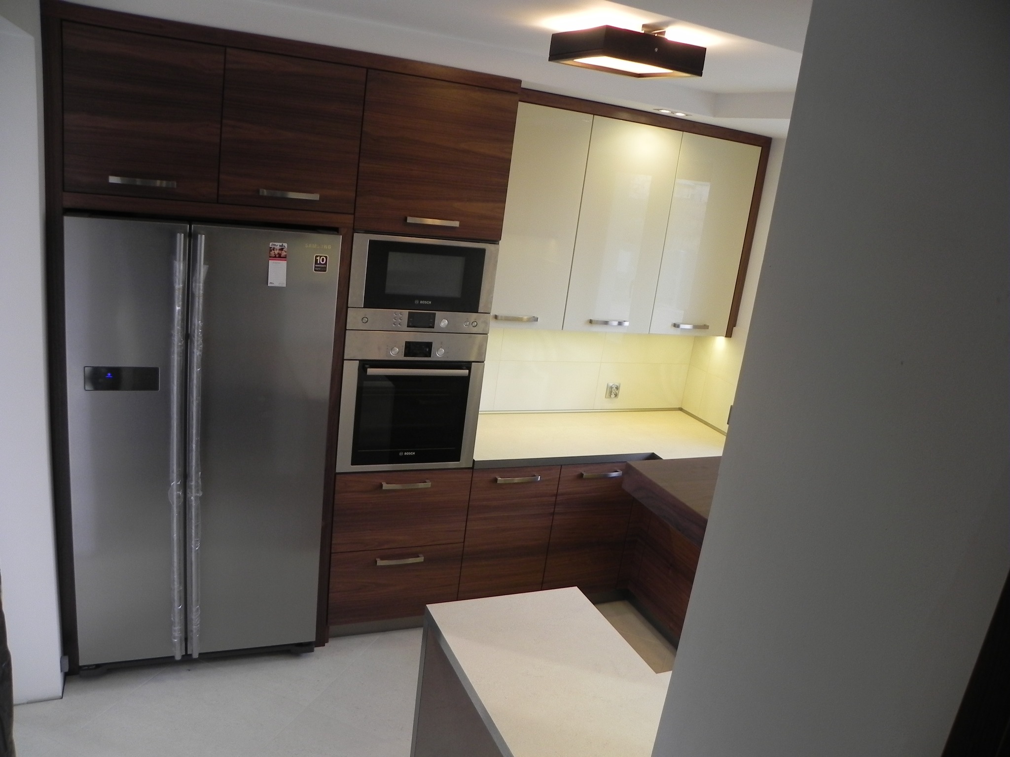 Nowoczesna kuchnia z ciemnego drewna i jasnymi szafkami, zabudowany sprzęt AGD: lodówka side-by-side, piekarnik i mikrofalówka marki Bosch. Elementy blatu z ciemnego drewna.