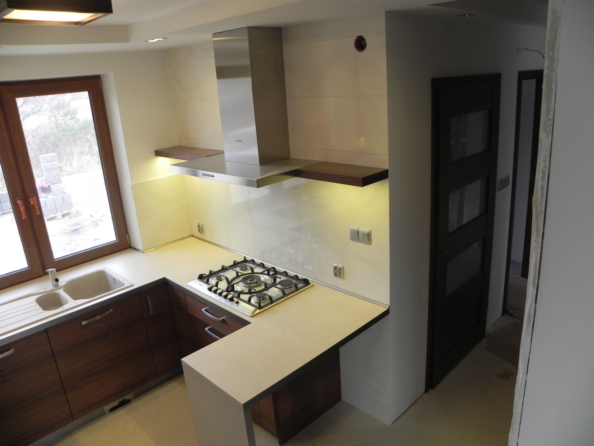 Nowoczesna kuchnia z brązowymi szafkami, jasnym blatem i płytą gazową; widoczny okap ze stali nierdzewnej i drewniane półki podświetlone od dołu. Przez okno widać stos bloczków betonowych.