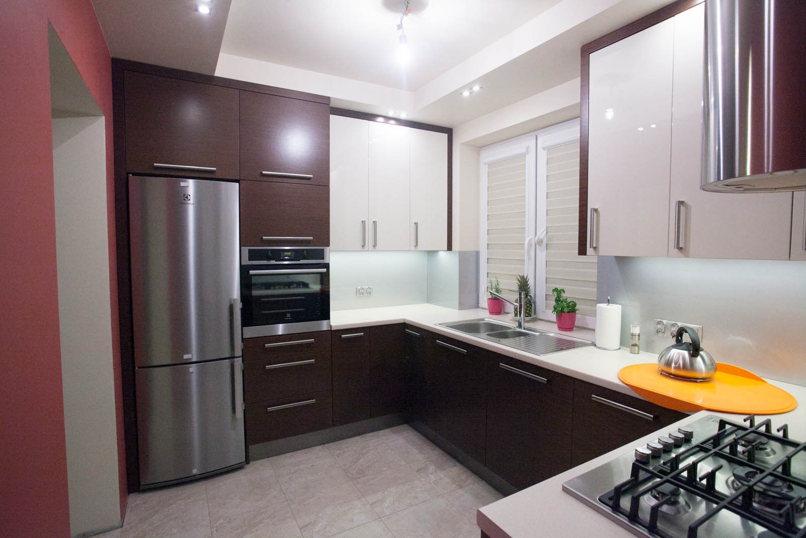 Nowoczesna kuchnia w brązach i bieli z chromowanymi akcentami, obejmująca zabudowę lodówki i piekarnika, z oświetleniem LED pod szafkami i roślinami doniczkowymi na blacie przy zlewie.