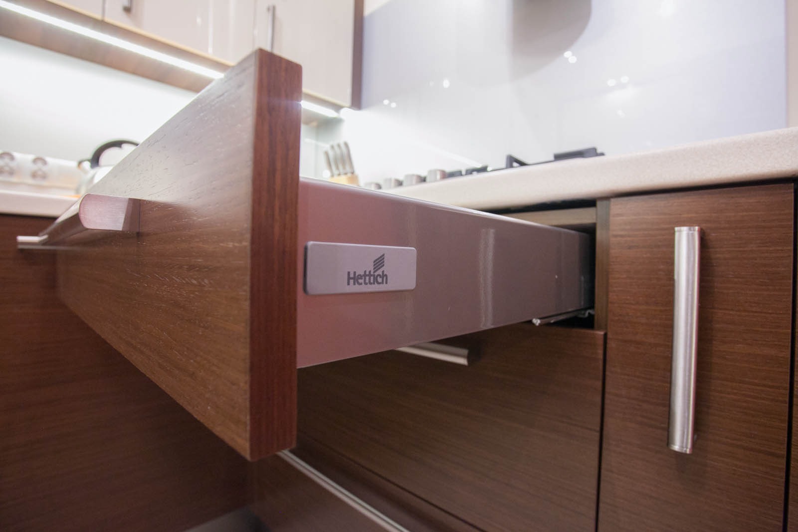 Wysuwana szuflada kuchenna z drewnianym frontem i systemem Hettich, widoczne oświetlenie podszafkowe i fragment blatu.