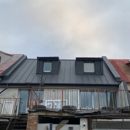 Zakład Ogólnobudowlany Dariusz Misiura - Szare, blaszane pokrycie dachowe z dwoma lukarnami na budynku z balkonem. Widoczne opierzenia blacharskie i fragment starego, czerwonego dachu sąsiedniego budynku.