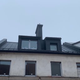 Zakład Ogólnobudowlany Dariusz Misiura - Nowe pokrycie dachowe z blachy na rąbek stojący, z oknami dachowymi i kominem, na tle pochmurnego nieba. Widoczna stara elewacja budynku.