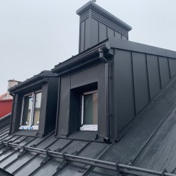 Zakład Ogólnobudowlany Dariusz Misiura - Nowoczesny, grafitowy dach z blachy na „click” z opierzeniami blacharskimi, widoczne okna dachowe i system odwadniania, w tle budynek z czerwoną dachówką.