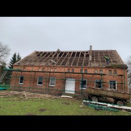 Zakład Ogólnobudowlany Dariusz Misiura - Ceglany budynek w trakcie prac dekarskich, widoczne rusztowania, częściowo zdjęte stare pokrycie i odsłonięta więźba dachowa, pracownicy na dachu, przyczepa z gruzem na trawniku.