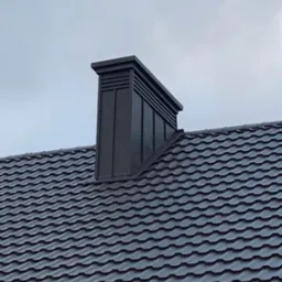 Szary dach z dachówki ceramicznej z nowoczesnym, metalowym kominem wentylacyjnym na tle pochmurnego nieba.