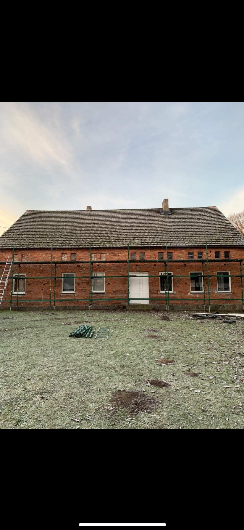 Ceglany budynek z szarym dachem, otoczony zielonymi rusztowaniami i zardzewiałą drabiną, na tle pochmurnego nieba, z fragmentem trawnika z widocznymi kretowiskami i stertą elementów rusztowania.