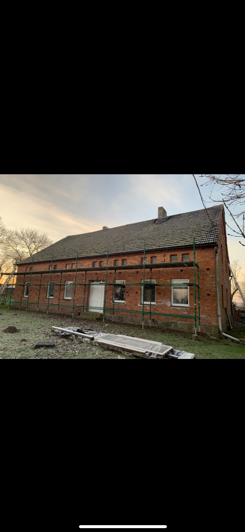 Ceglany budynek ze starym dachem, otoczony rusztowaniem podczas prac remontowych, widoczne elementy demontażu dachu na ziemi.