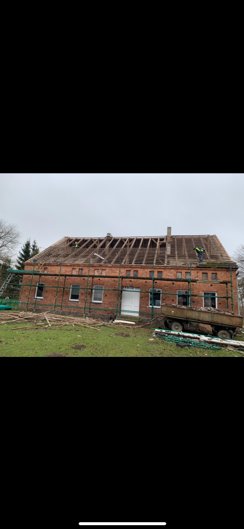 Ceglany budynek w trakcie prac dekarskich, widoczne rusztowania, częściowo zdjęte stare pokrycie i odsłonięta więźba dachowa, pracownicy na dachu, przyczepa z gruzem na trawniku.