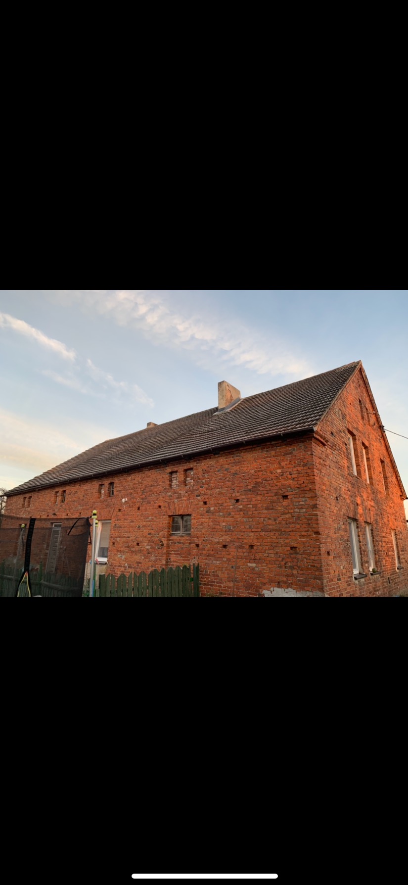 Ceglany budynek gospodarczy z dachem z ciemnej dachówki, widoczne ślady napraw i ubytki w murze.