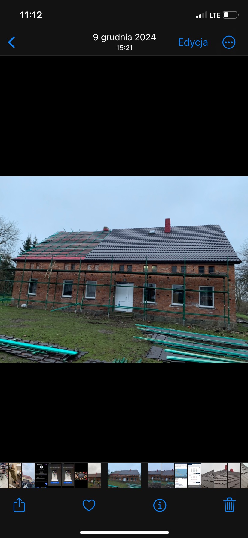 Częściowo wymieniony dach z ciemnej dachówki na ceglanym budynku gospodarczym, rusztowania wokół, przygotowane elementy dachowe na trawie.