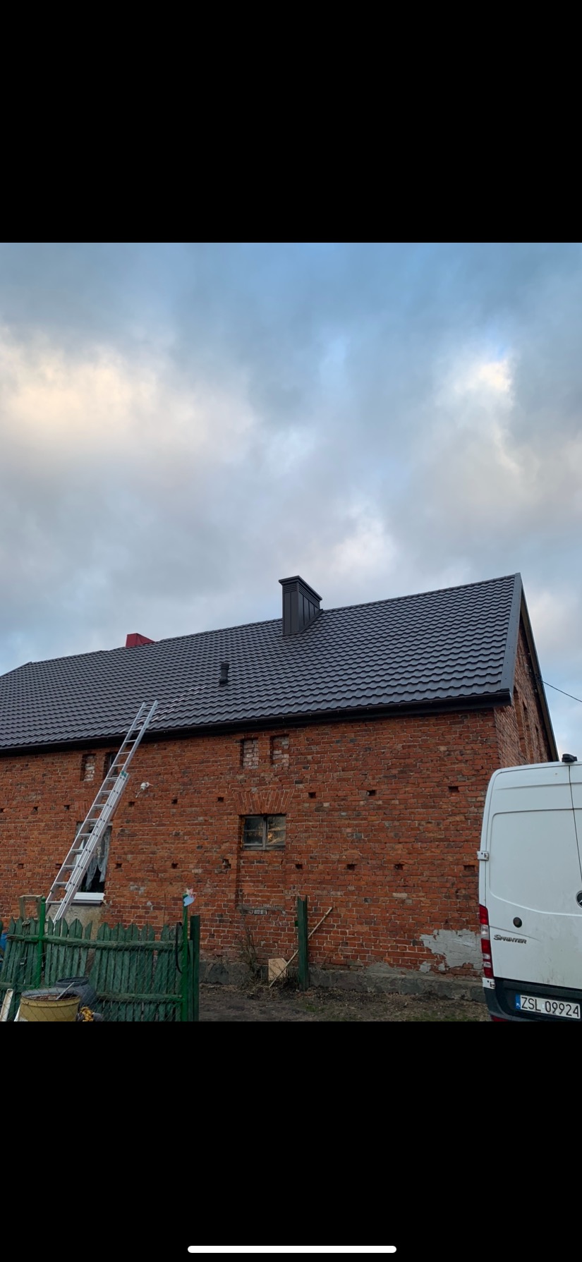 Ceglany budynek z nowym, ciemnoszarym dachem, na którym stoi drabina; widoczny fragment białego samochodu dostawczego oraz zielony płot.