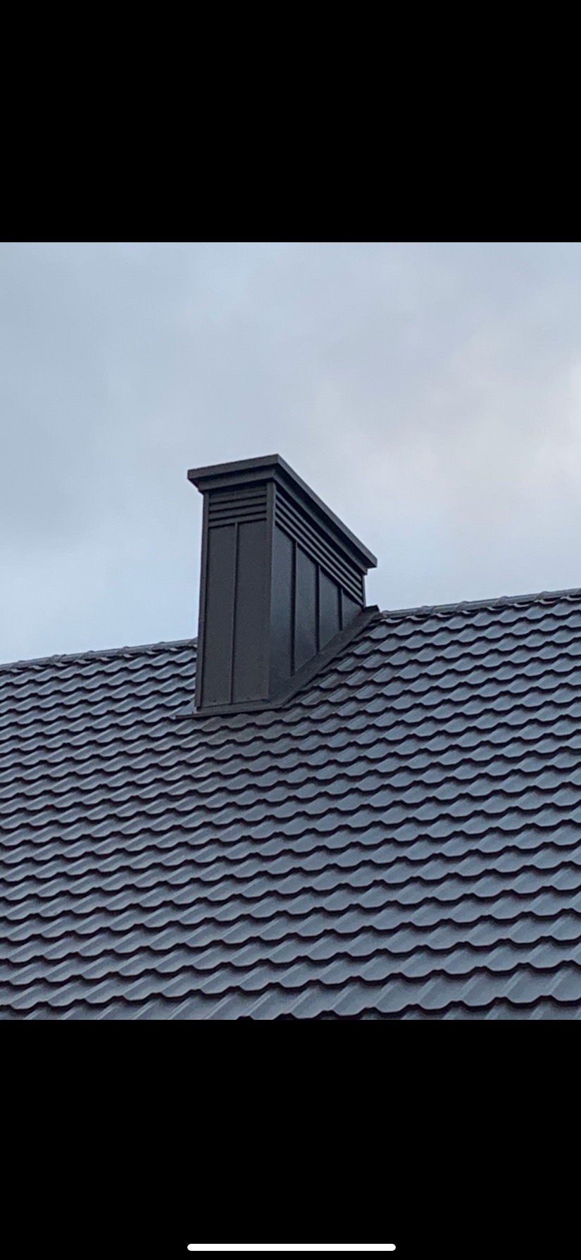 Szary dach z dachówki ceramicznej z nowoczesnym, metalowym kominem wentylacyjnym na tle pochmurnego nieba.