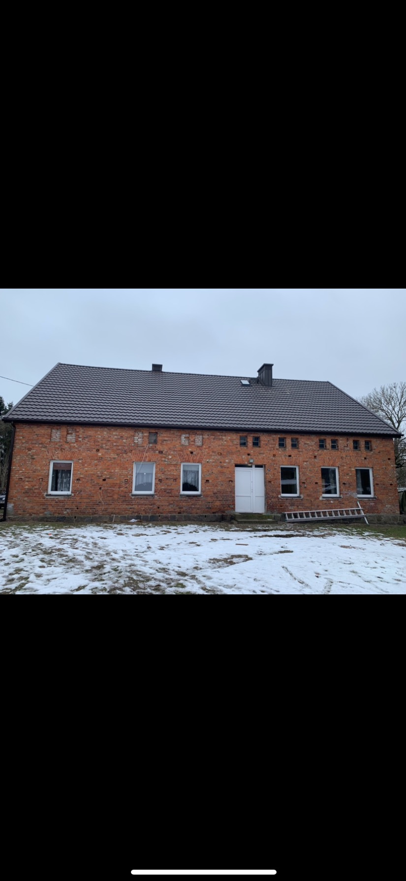 Ceglany budynek z nowym, brązowym dachem z dachówki, widoczne kominy i drabina oparta o ścianę, w tle zimowy krajobraz z resztkami śniegu.
