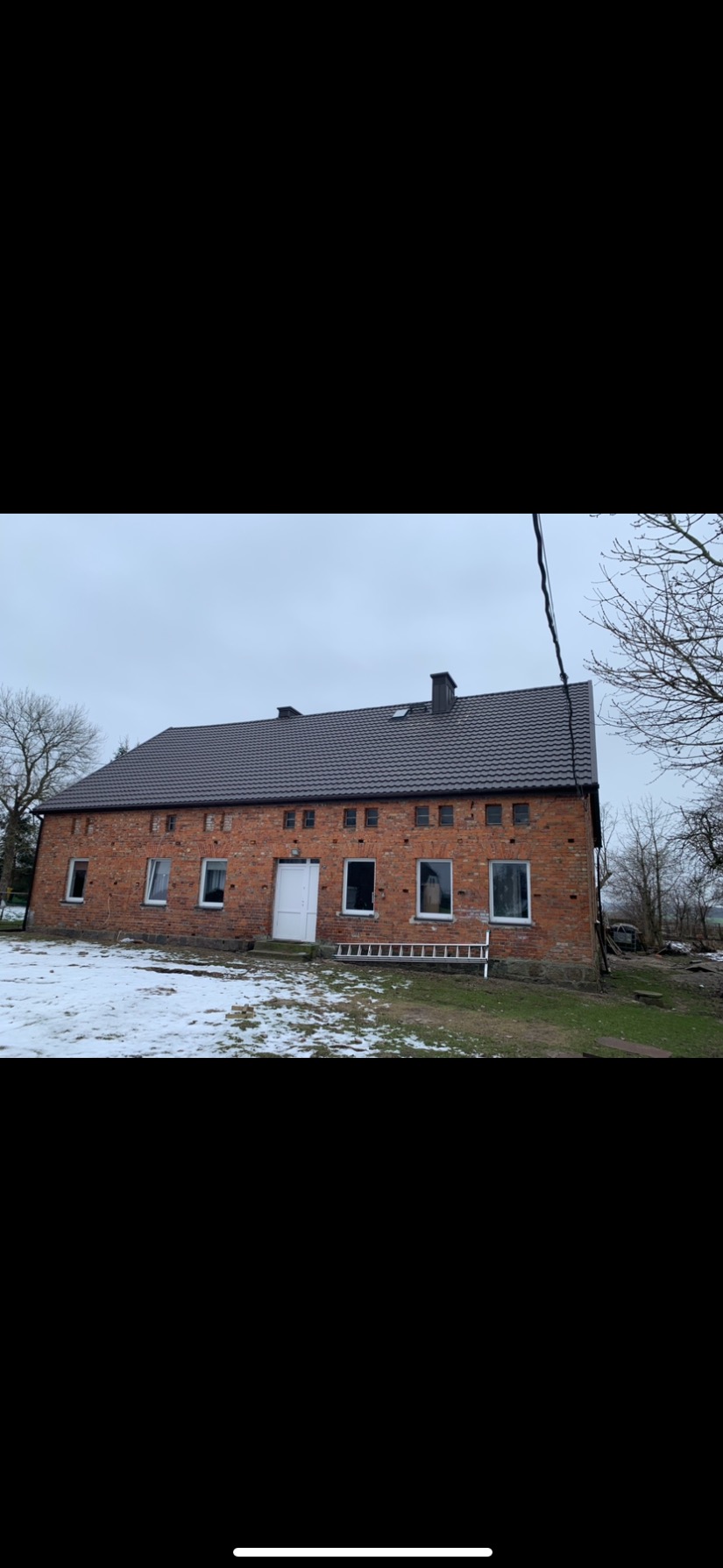 Dwukondygnacyjny budynek z czerwonej cegły z nowym, ciemnoszarym dachem, widoczna drabina oparta o ścianę, częściowo pokryty śniegiem teren.