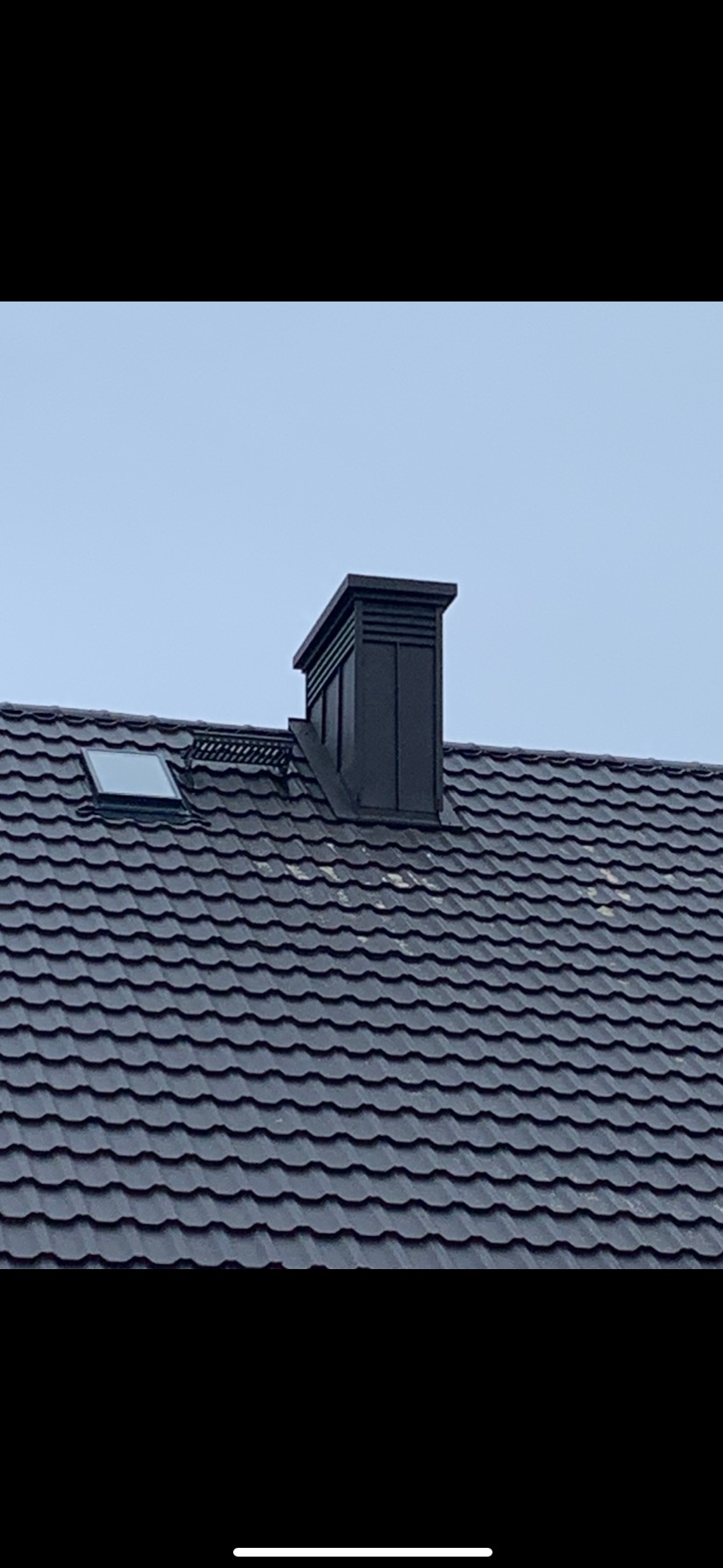 Czarny komin wentylacyjny na ciemnoszarym dachu z dachówki modułowej, widoczne okno dachowe