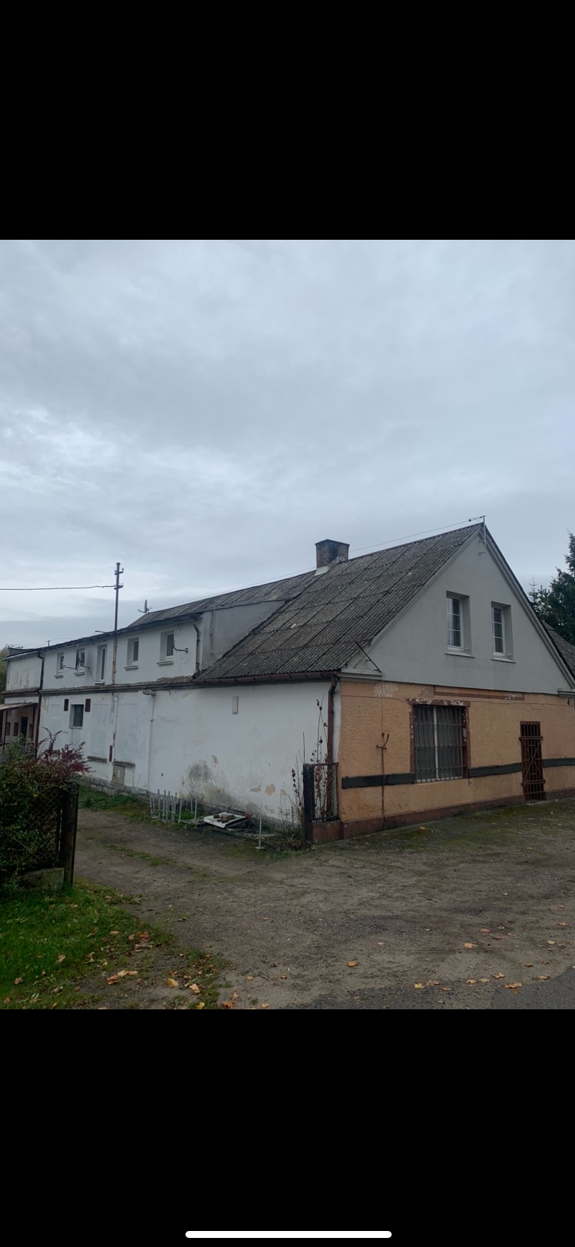 Widok zewnętrzny budynku z szarym, falistym dachem i częściowo odrapaną elewacją, sfotografowany w pochmurny dzień.