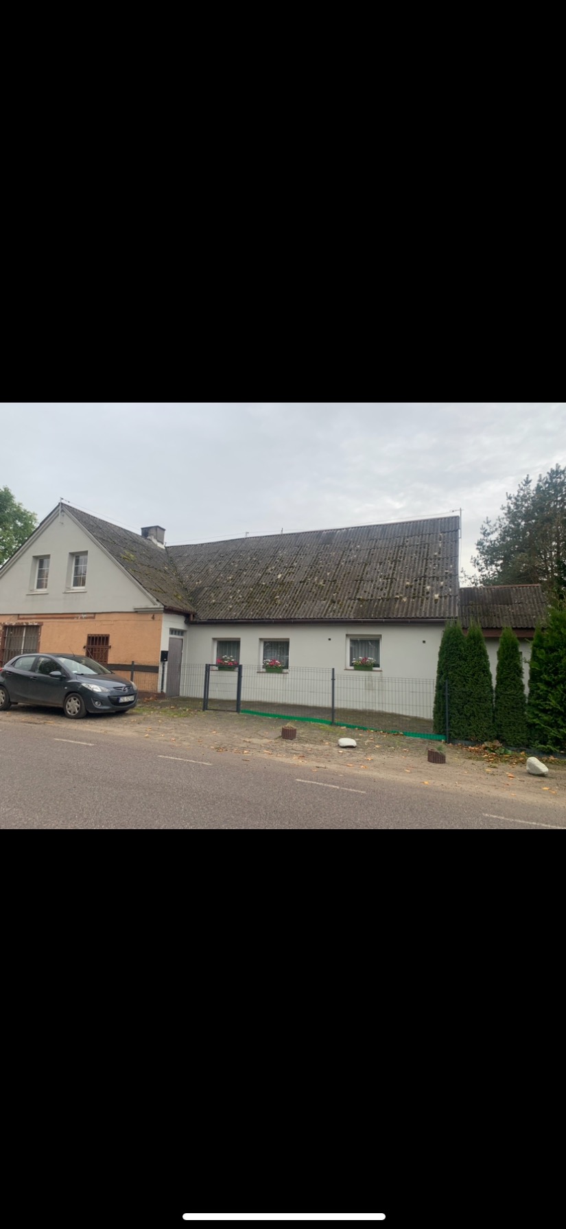 Dom jednorodzinny z szarym, lekko zarośniętym mchem dachem, widok z ulicy, szara Mazda zaparkowana przed budynkiem.