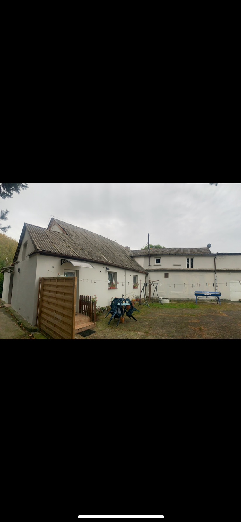 Dwukondygnacyjny budynek z szarym, lekko zabrudzonym dachem, widoczny z perspektywy podwórza z meblami ogrodowymi i huśtawką.