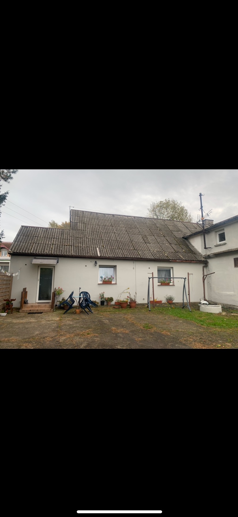 Dom jednorodzinny z szarym, lekko zabrudzonym dachem z eternitu, widok z podwórka z huśtawką i kwiatami w doniczkach.