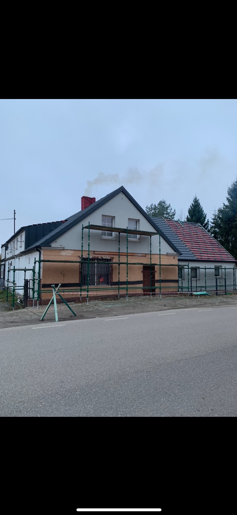 Dom jednorodzinny w trakcie prac dekarskich, widoczne rusztowania, część dachu pokryta nową dachówką, z komina unosi się dym.
