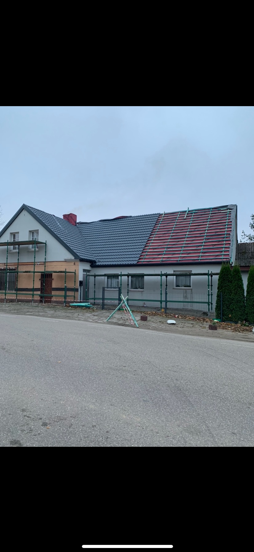 Dom w trakcie remontu dachu: część dachu pokryta szarą dachówką, druga część z widoczną konstrukcją i folią. Rusztowania wokół budynku, pochmurne niebo.