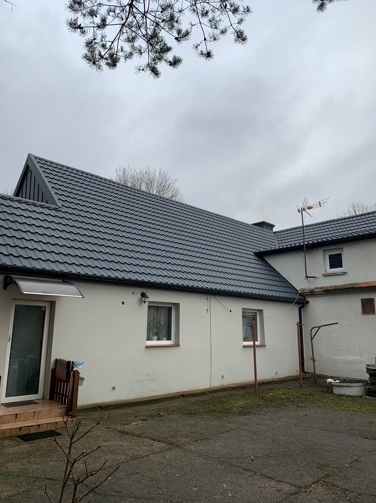 Dom jednorodzinny z nowym, ciemnoszarym dachem z blachy trapezowej, widoczny z podwórza w pochmurny dzień, antena telewizyjna na dachu budynku gospodarczego w tle.