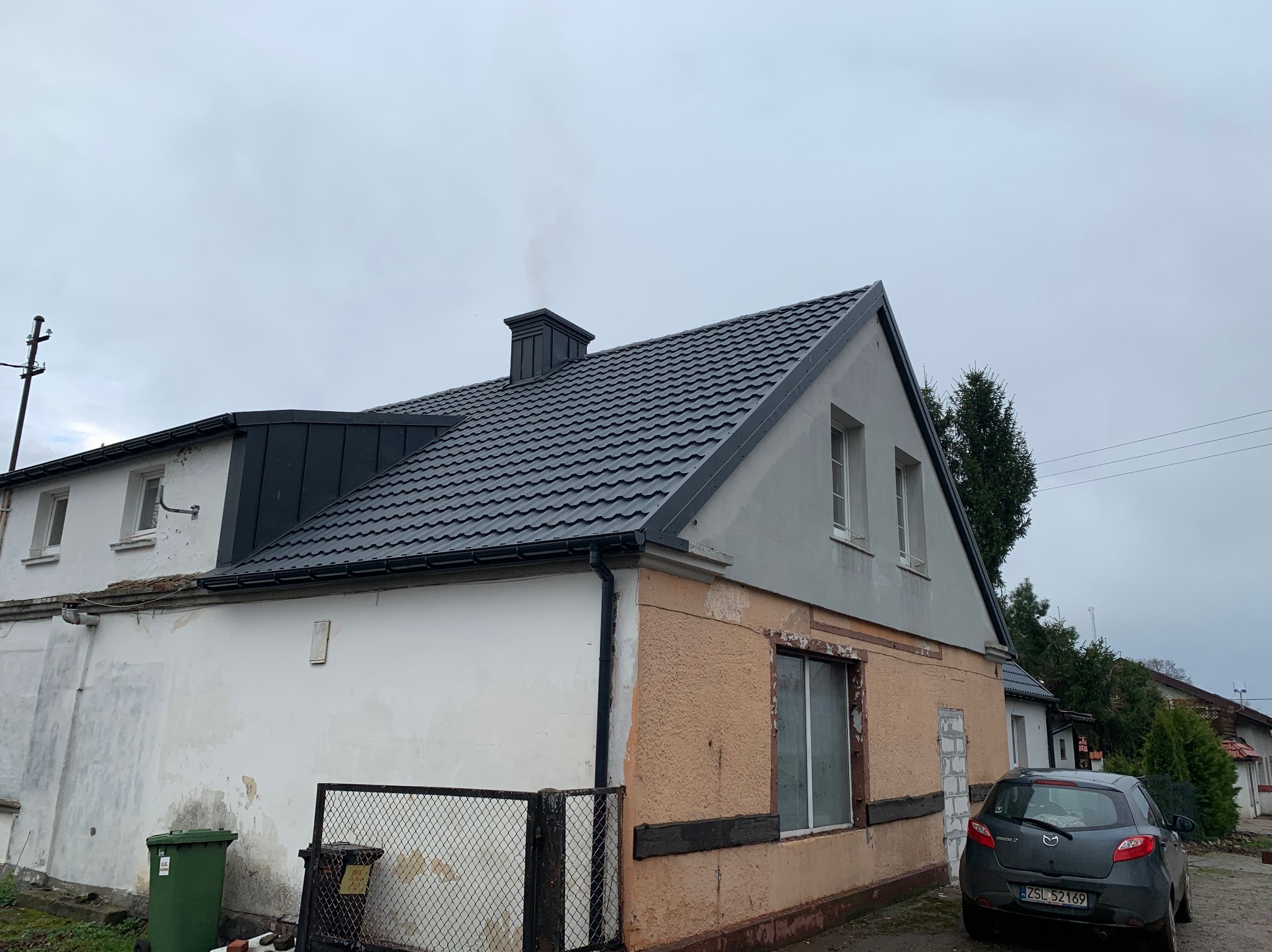 Dom z nowym, grafitowym dachem z dachówki ceramicznej i kominem, widok z boku, elewacja częściowo odnowiona, szare niebo.