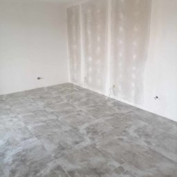 AK-BUD - Wnętrze pokoju z szarymi płytkami podłogowymi, ściany częściowo wyszpachlowane z wystającymi przewodami elektrycznymi.