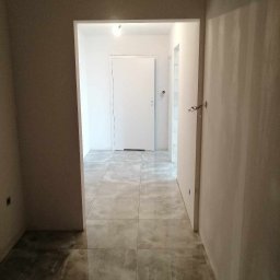 AK-BUD - Korytarz w trakcie remontu: świeżo położone, szare płytki podłogowe, ściany po szpachlowaniu, widoczne niedociągnięcia i wystające przewody elektryczne.