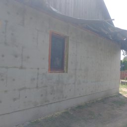 AK-BUD - Ściana budynku w trakcie ocieplania, widoczne płyty izolacyjne pokryte warstwą szpachli, fragment ściany z płyt OSB i instalacją elektryczną, okno z widoczną ramą i zabezpieczona góra budynku folią...