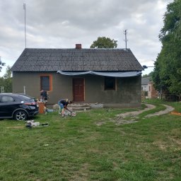 AK-BUD - Prace szpachlarskie na elewacji domu jednorodzinnego z szarą dachówką, widoczni dwaj pracownicy, narzędzia malarskie i samochód osobowy zaparkowany obok.