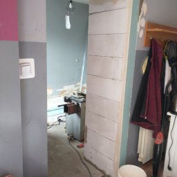 AK-BUD - Wnętrze w trakcie prac murarskich: ściana z białych bloczków, worki z zaprawą murarską NANO POZMUR-KL, widoczna maszyna do cięcia i wisząca lampa. Fragment ściany pomalowany na różowo i szaro.