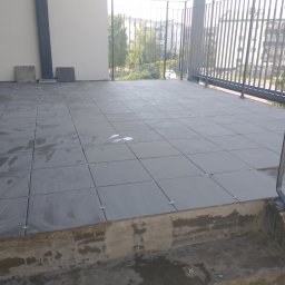 AK-BUD - Układanie szarych płytek na balkonie, widoczne krzyżyki dystansowe i ślady zaprawy, w tle balustrada i budynki mieszkalne.