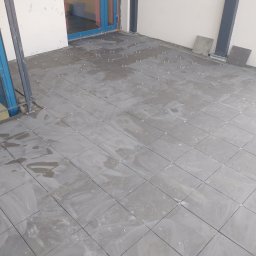 AK-BUD - Układanie szarych płytek na tarasie, widoczne krzyżyki dystansowe i resztki zaprawy, dwie płytki oparte o ścianę.