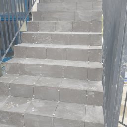 AK-BUD - Szare płytki na schodach zewnętrznych w trakcie układania, z widocznymi krzyżykami dystansowymi, metalowa balustrada obok.