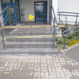 AK-BUD - Montaż płytek na schodach wejściowych do budynku z zabezpieczeniem drewnianym, widoczny worek zaprawy i roślinność przy barierce.