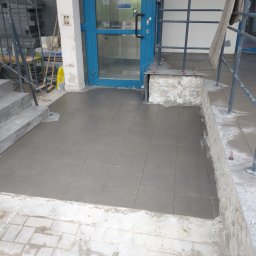 AK-BUD - Świeżo ułożona, szara, kwadratowa terakota na zewnątrz budynku, widoczne fugi i niedociągnięcia w wykończeniu krawędzi, niebieskie drzwi wejściowe w tle.